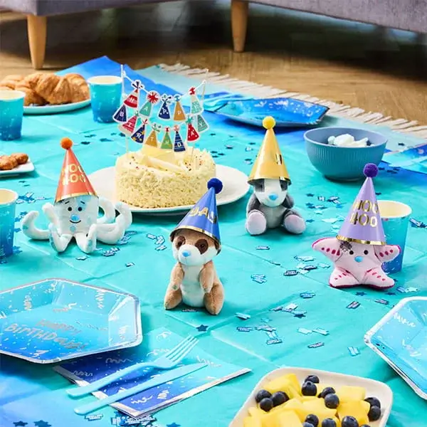 Blue birthday party table with a cake, party hats, sea-themed plush decorations, confetti, snacks, and themed tableware.